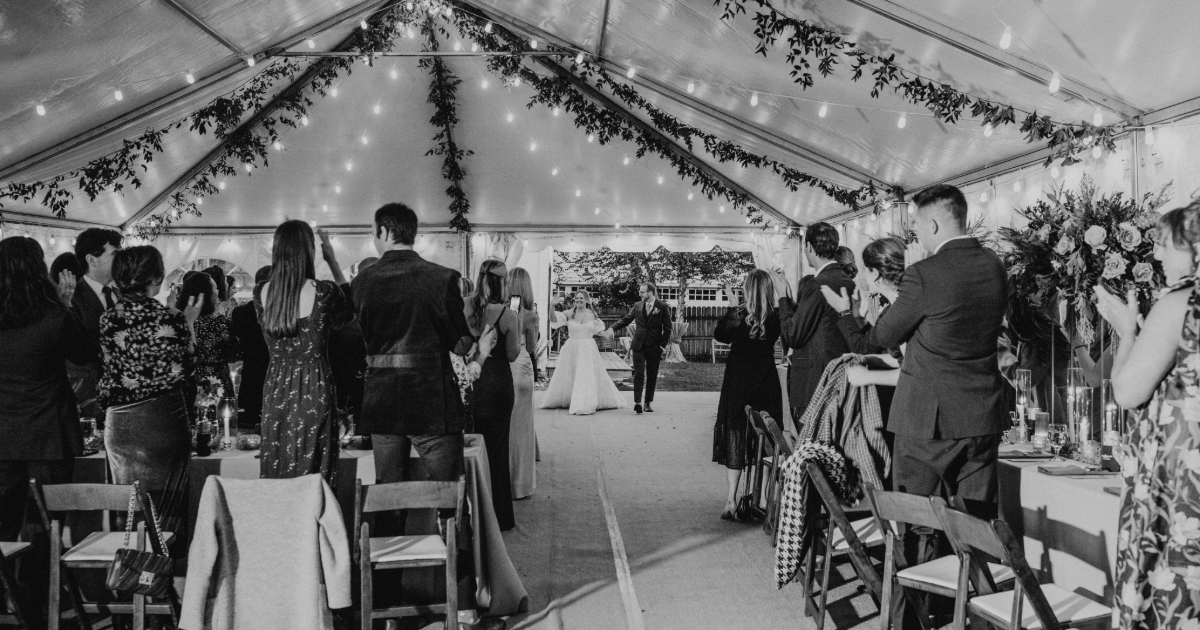 a bride and groom entering a tented wedding of guests standing and clapping