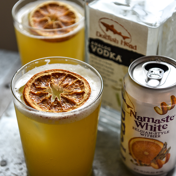 Glasses filled with Sun Salutation garnished with dried blood oranges next to a can of Namaste White and a bottle of Analog Vodka