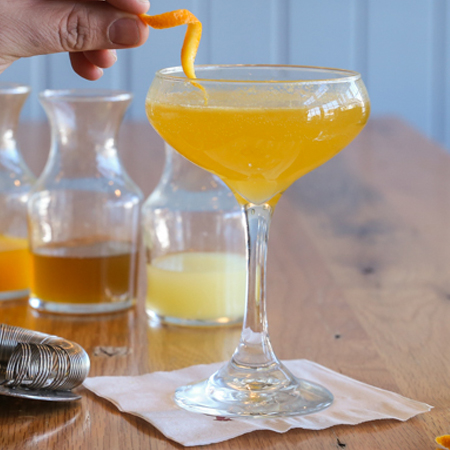 Cocktail glass filled with orange cocktail garnished with an orange peel surrounded by cocktail ingredients