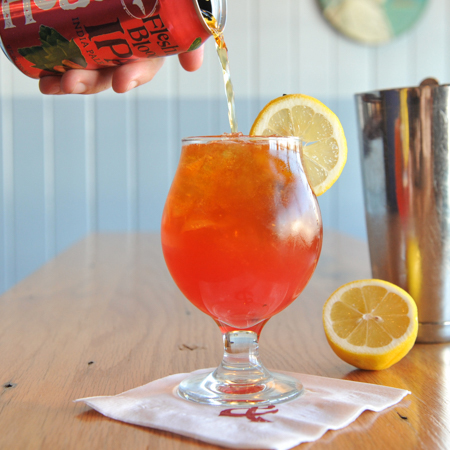 Person's hand pouring Flesh & Blood IPA into glass with cocktail garnished with lemon