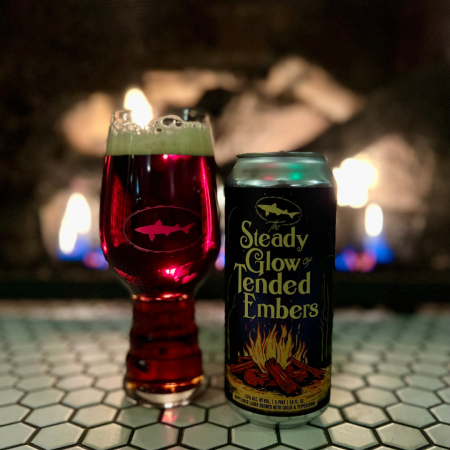 Glass pint of an amber-colored beer next to a 16 oz beer can that says "Steady Glow of Tended Embers" on it, with a fireplace burning behind the drinks