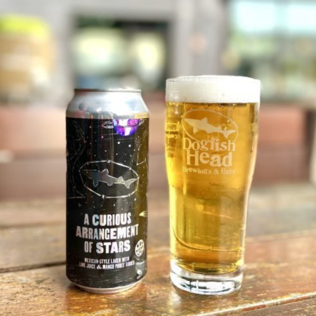 A can of 'A Curious Arrangement of Stars' on top of a table. To the right is a Dogfish Head pint glass filled with the Mexican-styled lager
