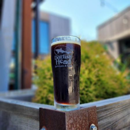 Dogfish Head beer Mother Nature Let’s Brew This that is Dark deep Amber Red with a tight tan head in a pint glass