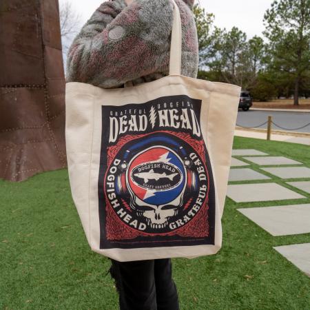 Front image of Dogfish Head x Grateful Dead tote bag. Large graphic with Grateful Dogfish Dead Head logo at top with red blue skull shark and shield art work. Slung on model's shoulder.
