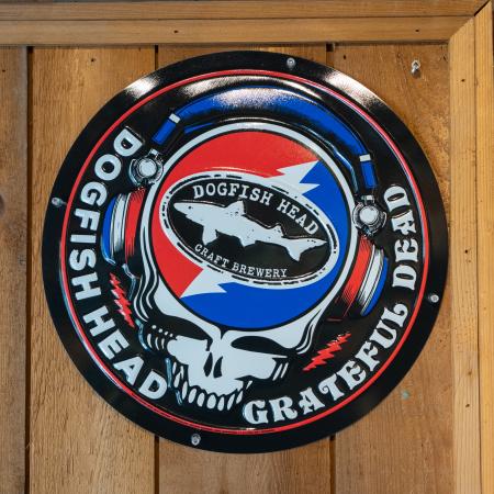 Circular metal wall tacker with the Dogfish Head x Grateful Dead skull with headphones and shark & shield logo 