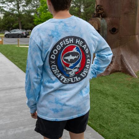 Person wearing a blue tie-dye long sleeve shirt with a large circular Dogfish Head x Grateful Dead graphic on the back featuring a shark logo within a red, white, and blue skull motif.