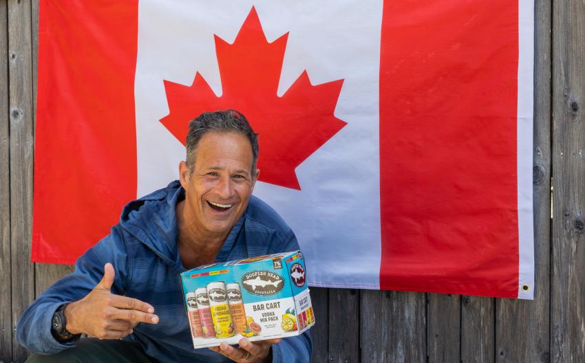 Sam Calagione holding a Bar Cart canned cocktail variety pack in front of a Canada flag