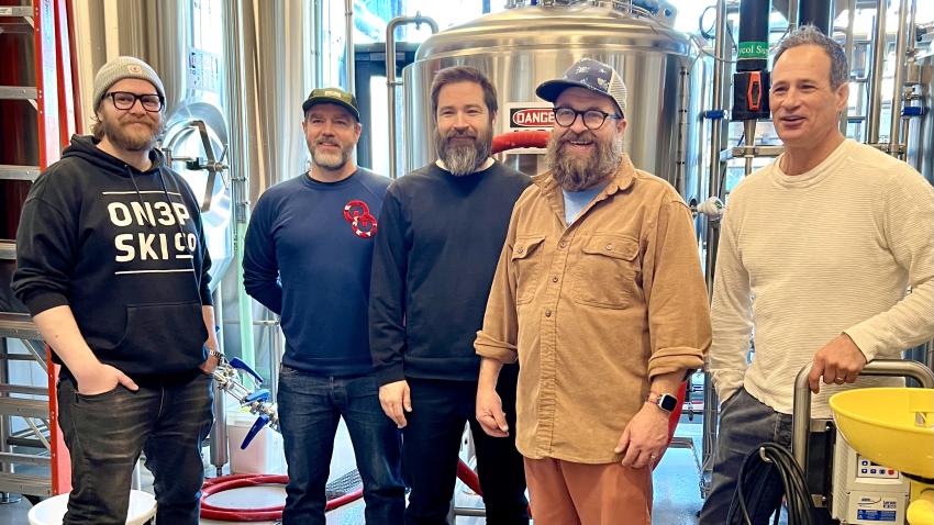 a group of brewers, five men, standing among silver brewing tanks