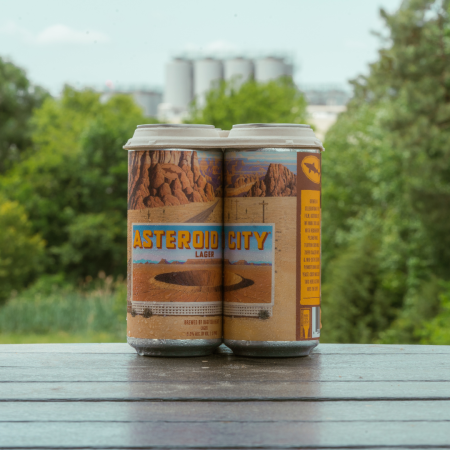 Four pack of 16 oz cans of Asteroid City Lager sitting atop a view with brewing towers in background