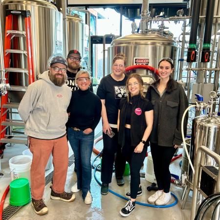 a group of 6 people posing together for a group photo with silver beer brewing equipment in the background
