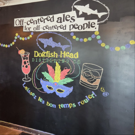 a chalkboard with a mardi gras mask on it in purples and yellows that says Dogfish Head