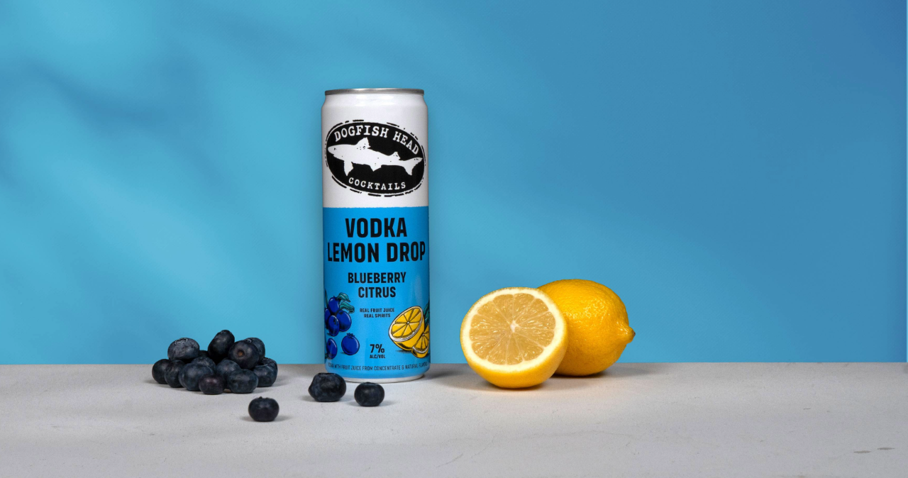 Dogfish Head Blueberry Citrus Vodka Lemon Drop on a table top with a blue background and lemons and blueberries around it