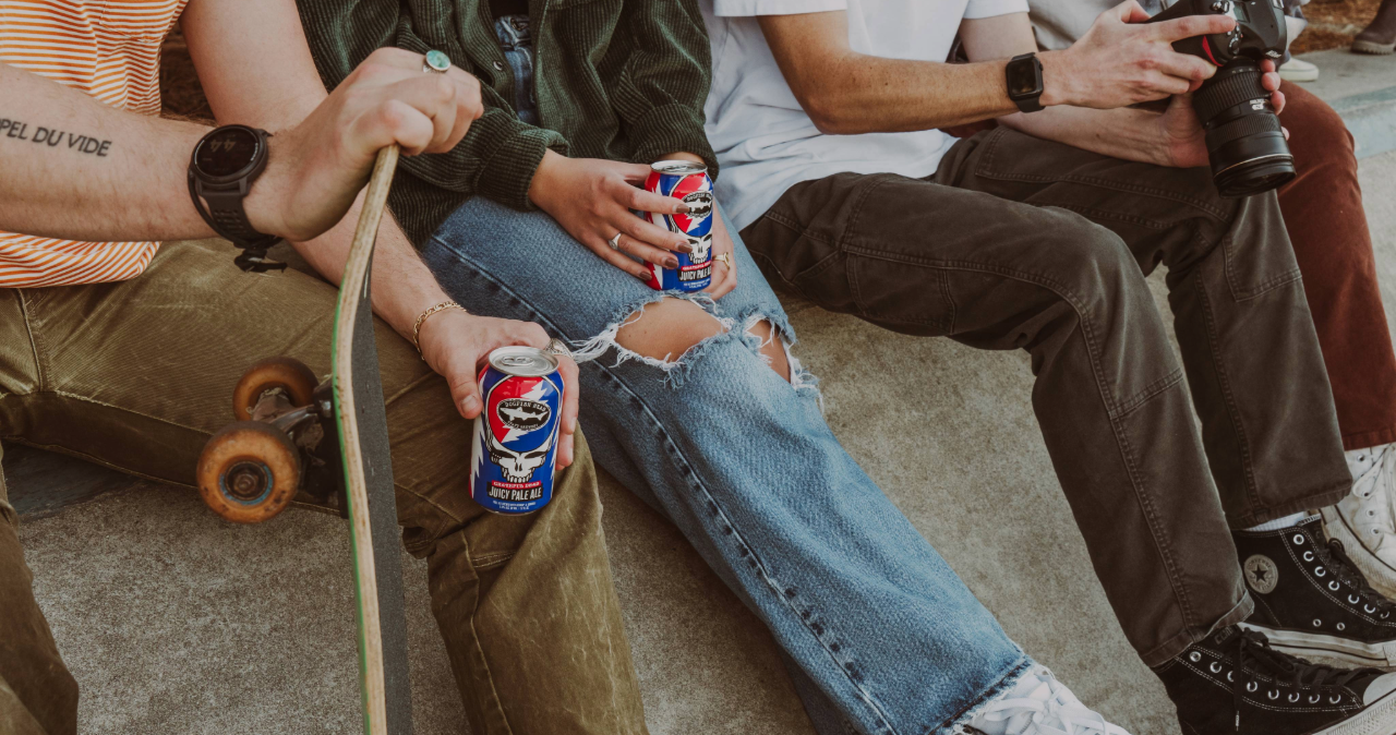 A few folks in a skate park with two holding 12 oz cans of Dogfish Head x Grateful Dead Juicy Pale Ale