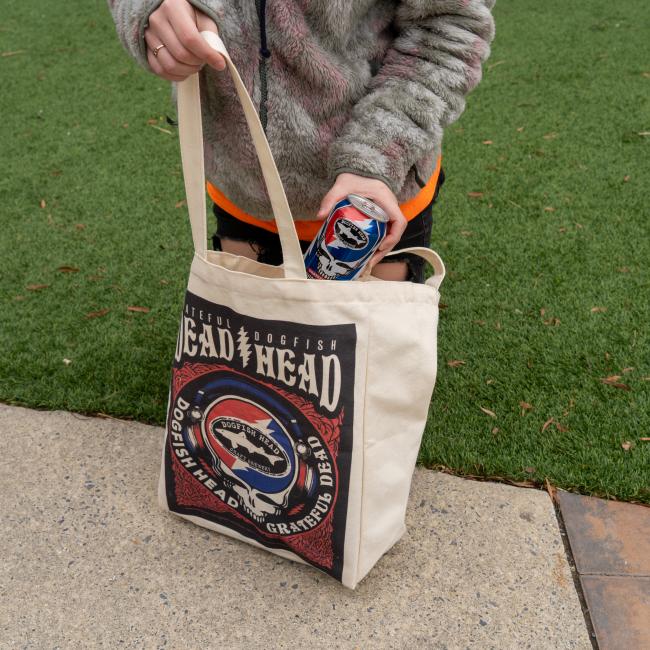 Front image of Dogfish Head x Grateful Dead tote bag. Large graphic with Grateful Dogfish Dead Head logo at top with red blue skull shark and shield art work. Featured with collaboration beer can. 