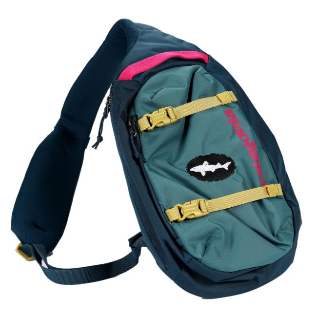 Angled view of the Patagonia sling backpack highlighting its contoured shape, padded shoulder strap, front patch with Dogfish Head shark logo, and dual yellow compression straps.