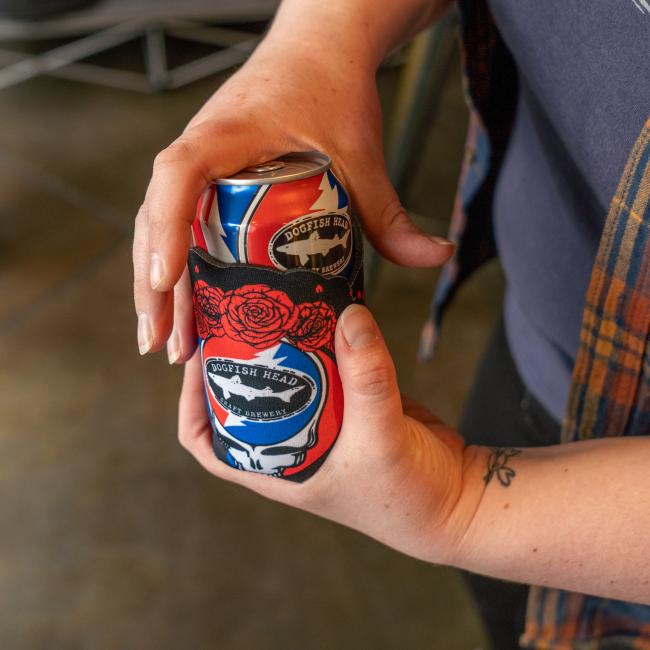 A black can cooler with the Dogfish Head and Grateful Dead collaboration skull logo on the front and red roses around the top. Pictured here being held for scale.