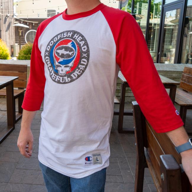 Person wearing a white and red raglan-style baseball tee featuring a circular Dogfish Head x Grateful Dead graphic on the chest.