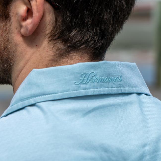 Close up of the back collar of blue button up short sleeve shirt. Embroidered with Howler logo in center.