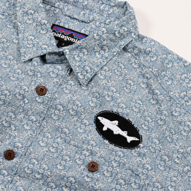 Close-up of the shirt’s upper front section showing the fine blue and white pattern, Patagonia brand tag inside the collar, and an embroidered black and white Dogfish Head shark logo patch on the chest.