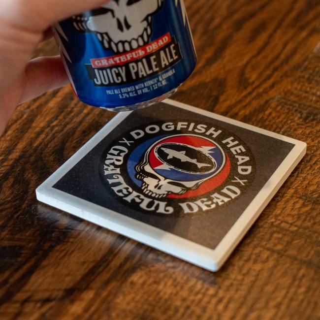 Square ceramic coaster featuring Dogfish x Grateful Dead collaboration artwork with skull, shark and shield. Shot next to collaboraion can. 