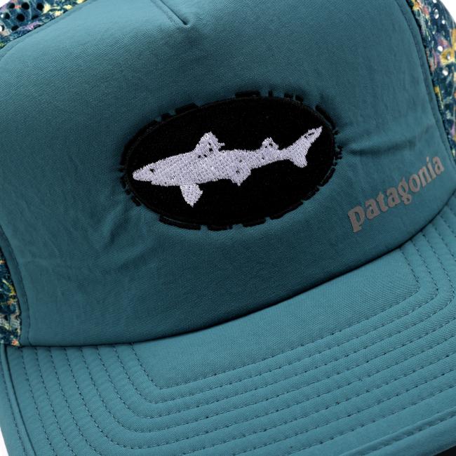 Close-up of the front panel of the hat, showcasing the black patch with a stitched white shark silhouette and the light gray Patagonia branding on the teal fabric.
