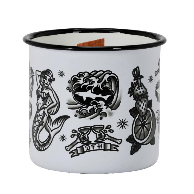A white enamel mug candle featuring vintage-style black-and-white tattoo graphics, including anchors, mermaids, citrus, and Dogfish Head branding, with a wooden wick in the center.