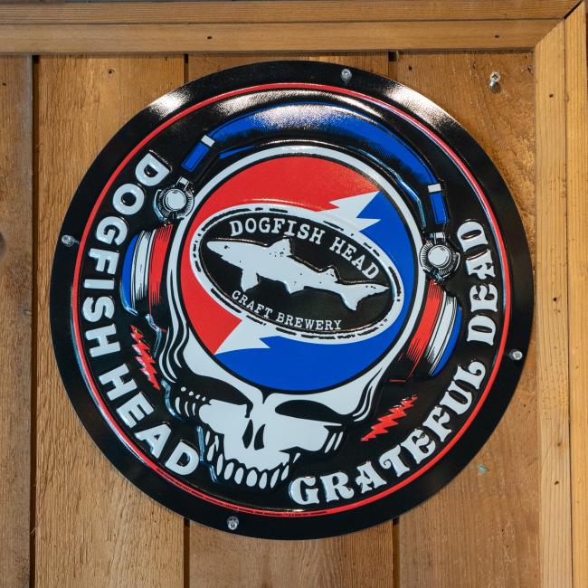 Circular metal wall tacker with the Dogfish Head x Grateful Dead skull with headphones and shark & shield logo 