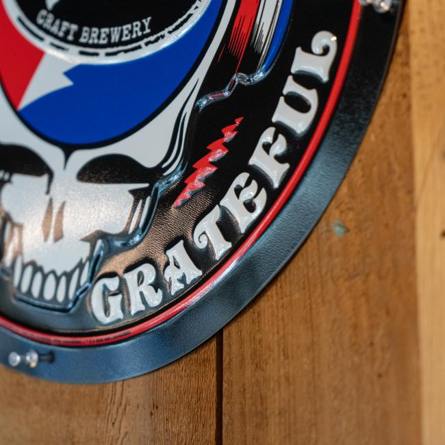 Circular metal wall tacker with the Dogfish Head x Grateful Dead skull with headphones and shark & shield logo 
