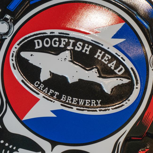 Circular metal wall tacker with the Dogfish Head x Grateful Dead skull with headphones and shark & shield logo 