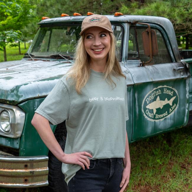 A smiling person poses in front of a weathered Dogfish Head-branded green pickup truck while wearing the Beer and Benevolence t-shirt and matching tan hat.