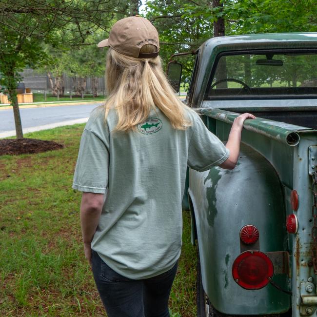 Rear view of the person wearing the full Beer and Benevolence outfit, showing both the back of the t-shirt and the hat with matching logos, standing beside the green truck.